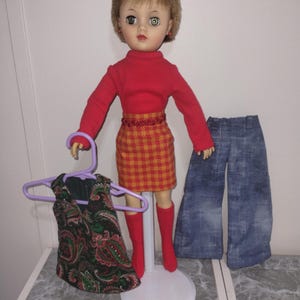 May include: A vintage doll with short blonde hair, wearing a red turtleneck, a plaid skirt, and red boots. A paisley print top hangs on a purple hanger. Blue denim bell-bottom pants are also included.