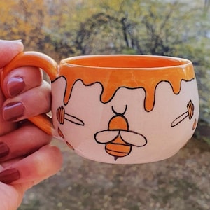 May include: A white ceramic mug with a honey-dripping design and a bee illustration. The mug is painted with orange and yellow accents.