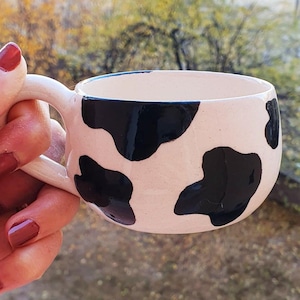 May include: A white ceramic mug with black cow print design. The mug has a handle and is held by a hand.