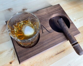 May include: A wooden cigar and whiskey set. The set includes a dark brown wooden tray with a circular cutout for a cigar and a round cutout for a glass. A glass of whiskey with a large ice cube sits next to the cigar. The letter "A" is carved into the wood.