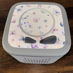 May include: A grey speaker with a white top decorated with a butterfly pattern in pink, blue, and purple. The speaker has a perforated front and two dark grey control buttons.