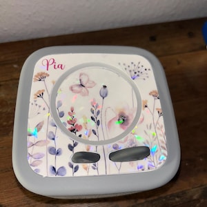 Könnte beinhalten: Ein grauer, quadratischer Behälter mit floralem Design und dem Namen "Pia" in rosa Schrift. Der Deckel hat einen weißen Hintergrund mit Aquarellblumen, einem Schmetterling und schillernden Akzenten. Der Behälter steht auf einer Holzoberfläche.
