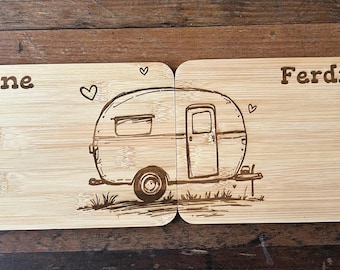 Könnte beinhalten: Zwei Holzschneidebretter mit einer Wohnwagenillustration und den eingravierten Namen "Simone" und "Ferdinand". Der Wohnwagen hat eine Tür, ein Fenster und ein Rad. Herzen sind in der Nähe des Wohnwagens und der Namen geätzt.