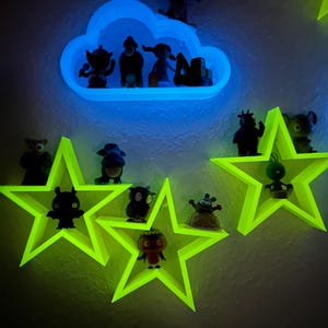 May include: A wall display with neon green star and blue cloud-shaped shelves, holding small figurines. The shelves glow due to the lighting. The figurines are various shapes and sizes. The wall is painted a neutral colour, providing a contrasting backdrop.