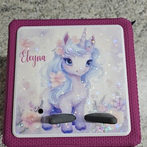 Könnte beinhalten: Ein magentafarbenes, quadratisches Gerät mit weißem Rahmen. Das Gerät zeigt eine Einhorn-Illustration mit blauem Haar, einem Horn und floralen Akzenten. Der Name "Eleyna" ist kursiv geschrieben. Zwei schwarze Knöpfe sind sichtbar.