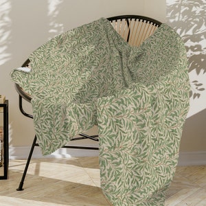 May include: A green and cream throw blanket with a leafy pattern, draped over a wicker chair in a modern living room setting.