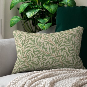 May include: A decorative throw pillow with a green and white willow pattern. The pillow is on a light gray couch with a white throw blanket.