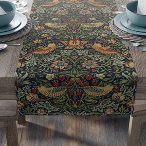 May include: A dark blue table runner with a repeating pattern of birds, flowers, and leaves. The pattern is in shades of green, yellow, orange, and red. The runner is on a wooden table with two place settings on each side.
