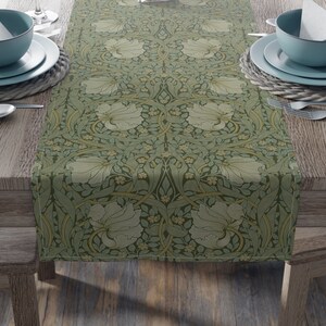 May include: A green table runner with a floral pattern featuring white flowers and yellow accents. The runner is on a wooden table with two place settings on each side.