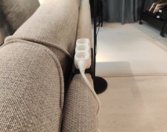 May include: A white three-outlet power strip attached to the side of a beige couch.