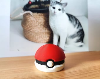 May include: A red and white Poke Ball toy with a black band around the middle. The Poke Ball is sitting on a wooden surface.