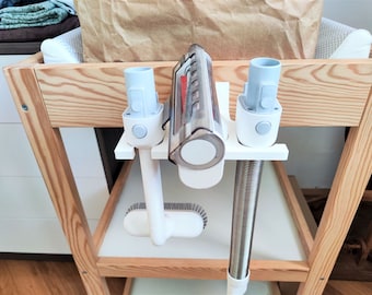 May include: A white vacuum cleaner attachment organizer with three attachments hanging from it. The organizer is mounted on a wooden shelf. The attachments include a crevice tool, a dusting brush, and a hose.