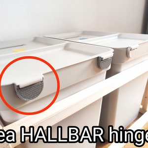 Può includere: Due contenitori di plastica bianca con cerniere grigie. I contenitori sono impilati su una struttura in legno. I contenitori sono etichettati con "IKEA HALLBAR hinges".