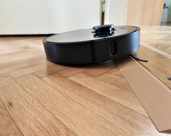 May include: A black robotic vacuum cleaner with a round, reflective top and a black bumper. The vacuum is on a wooden floor with a brown strip of tape.