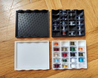 May include: A white plastic storage box with 16 compartments for storing sewing thread spools. The box is empty and has a wavy edge. A black plastic storage box with 16 compartments for storing sewing thread spools is also shown. The box is filled with colorful thread spools.