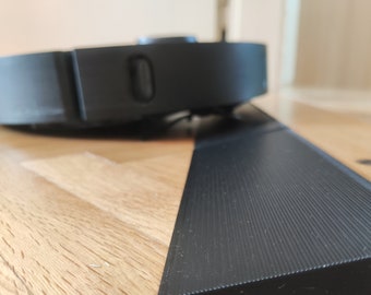 May include: A black robot vacuum cleaner with a textured surface and a small button on the side. The vacuum cleaner is sitting on a wooden floor.