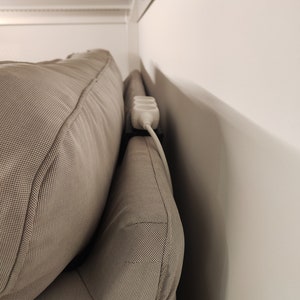 Könnte beinhalten: Eine weiße Steckdosenleiste mit einem schwarzen Kabel ist hinter einem beigen Sofa versteckt.