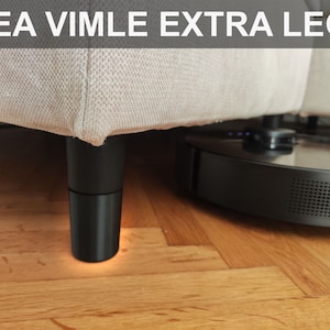 Op de afbeelding: Zwarte meubelpoten ondersteunen een beige bank. De afbeelding toont de onderkant van de bank met de tekst "IKEA VIMLE EXTRA LEGS" bovenaan. Een zwarte robotstofzuiger is zichtbaar onder de bank.