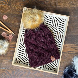 May include: A burgundy cable-knit beanie with a faux fur pom-pom. The hat is displayed on a wooden tray with a geometric design. The pom-pom is a mix of brown and tan colours. A small leather tag is sewn on the hat.