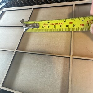 K&ouml;nnte beinhalten: Ein Holzrahmen mit neun quadratischen F&auml;chern. Ein gelbes Ma&szlig;band ist oben auf dem Rahmen platziert und zeigt eine Messung von 25,4 cm.