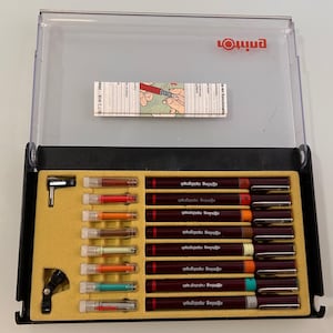 May include: A black case containing a set of Rotring Rapidograph technical pens. The pens are dark red with black caps and various colored tips. The case also includes a small wrench and a small black ink bottle.