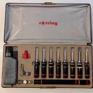 May include: A black hardshell carrying case containing a set of eight technical pens with black barrels and colored rings, a black ink bottle labeled rotring, a metal compass attachment, and a long black pen holder with red and yellow bands.