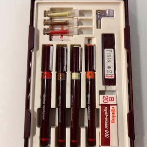 Op de afbeelding: Een bordeauxrode plastic etui met vier Rotring Rapidograph technische pennen met gekleurde ringen, een metalen passerhulpstuk, een doosje met 0,5 HB vullingen en een Rotring B20 rapid-eraser, geplaatst in voorgevormde vakjes.