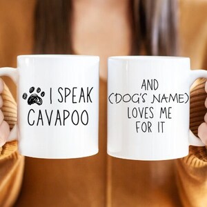 Op de afbeelding: Twee witte koffiemokken met zwarte tekst. De mok aan de linkerkant zegt "Ik spreek Cavapoo" met een pootprint. De mok aan de rechterkant zegt "En (Naam van de hond) houdt van me ervoor".