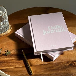Może przedstawiać: Dwa r&oacute;żowe dzienniki, ułożone jeden na drugim na drewnianej powierzchni. G&oacute;rny dziennik ma napis "Daily Journal" wydrukowany na okładce na biało.