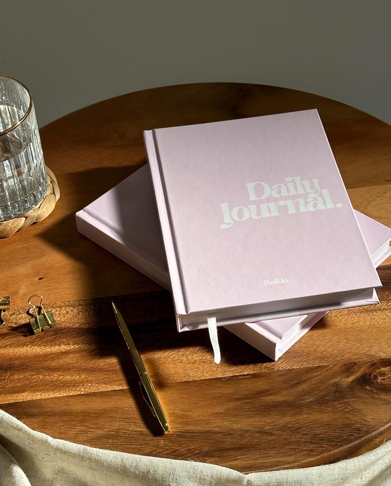 Może przedstawiać: Dwa r&oacute;żowe dzienniki, ułożone jeden na drugim na drewnianej powierzchni. G&oacute;rny dziennik ma napis "Daily Journal" wydrukowany na okładce na biało.