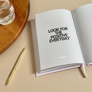Może przedstawiać: Biały dziennik z nadrukowanym czarnym tekstem "LOOK FOR THE POSITIVE EVERYDAY". Dziennik jest otwarty na pustej stronie i ma r&oacute;żową wstążkę jako zakładkę. Dziennik leży na jasnobrązowej powierzchni z szklanką wody i złotym długopisem.