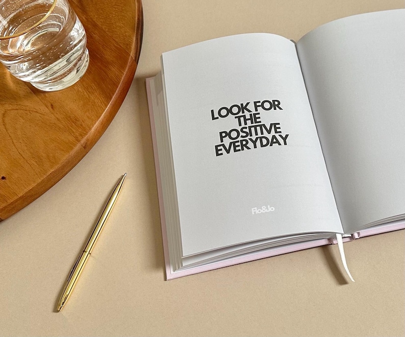 Może przedstawiać: Biały dziennik z nadrukowanym czarnym tekstem "LOOK FOR THE POSITIVE EVERYDAY". Dziennik jest otwarty na pustej stronie i ma r&oacute;żową wstążkę jako zakładkę. Dziennik leży na jasnobrązowej powierzchni z szklanką wody i złotym długopisem.