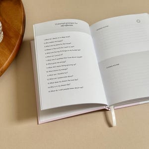 Może przedstawiać: Otwarty dziennik w r&oacute;żowej okładce z białymi stronami. Lewa strona zawiera 15 podpowiedzi do dziennika do refleksji nad sobą. Prawa strona zawiera sekcje na poranne i wieczorne rutyny.
