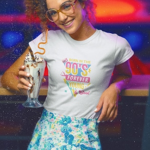 Op de afbeelding: Wit T-shirt met een kleurrijke jaren 90-afbeelding met de tekst "Born in the 90's Forever Young & Cool". De persoon draagt een bloemenrok en houdt een milkshake vast.