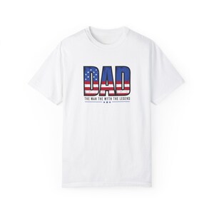 Op de afbeelding: Wit T-shirt met het woord "DAD" in een versleten, patriottisch ontwerp. De letters zijn gevuld met het Amerikaanse vlagpatroon. Onder het woord "DAD" staat de tekst "THE MAN THE MYTH THE LEGEND" met drie sterren.