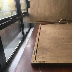 May include: Two wooden trays with a light brown finish. One tray has a woven handle. The trays are rectangular with a decorative leaf design in the corners. The trays are on a brown surface near a window.