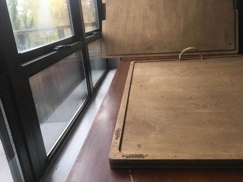 May include: Two wooden trays with a light brown finish. One tray has a woven handle. The trays are rectangular with a decorative leaf design in the corners. The trays are on a brown surface near a window.