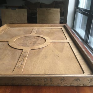 May include: A large, rectangular wooden tray with a light brown finish. The tray features a central circular design and decorative carvings. The edges are framed with detailed patterns. The tray is placed on a wooden surface.