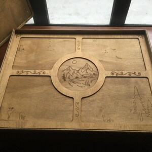 May include: Wooden tray with a landscape design. The tray features a central circular design with mountains, trees, and a sun. The corners and edges have decorative carvings of animals, trees, and swirls. The tray is made of light-coloured wood.