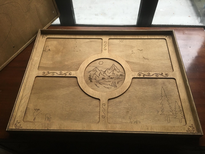 May include: Wooden tray with a landscape design. The tray features a central circular design with mountains, trees, and a sun. The corners and edges have decorative carvings of animals, trees, and swirls. The tray is made of light-coloured wood.