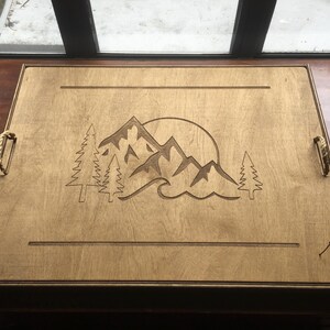 May include: Wooden serving tray with a mountain and sun design. The rectangular tray features rope handles and a laser-etched mountain scene with trees and a sun. The wood has a light brown colour.