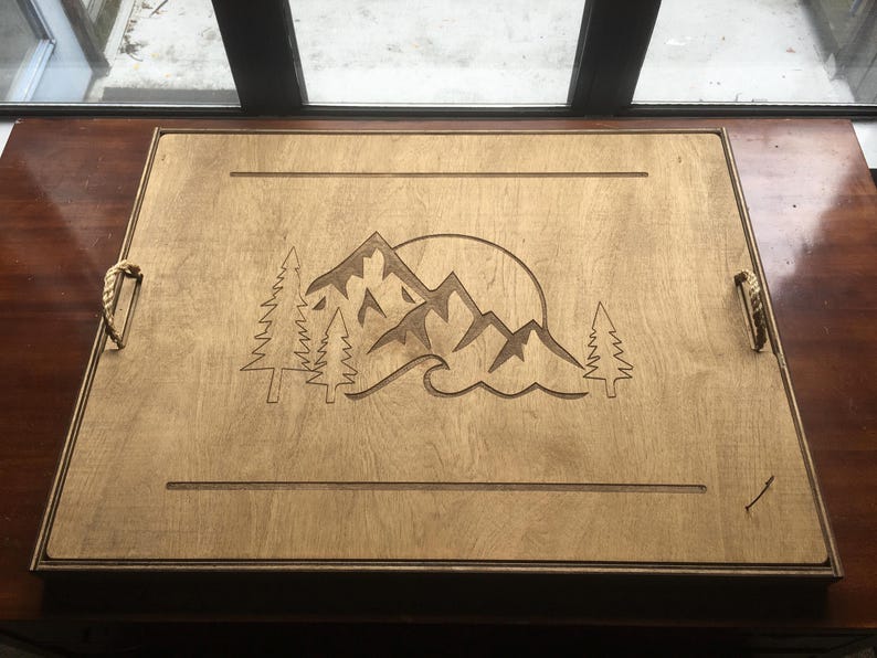 May include: Wooden serving tray with a mountain and sun design. The rectangular tray features rope handles and a laser-etched mountain scene with trees and a sun. The wood has a light brown colour.