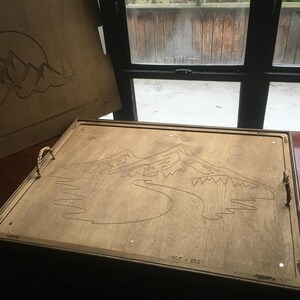 May include: Wooden serving tray with a mountain and sun design. The tray has rope handles and measures 77.5 x 58.4 cm. The design is etched into the wood, creating a rustic aesthetic.