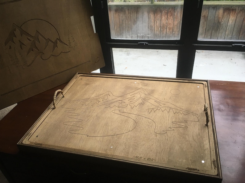 May include: Wooden serving tray with a mountain and sun design. The tray has rope handles and measures 77.5 x 58.4 cm. The design is etched into the wood, creating a rustic aesthetic.