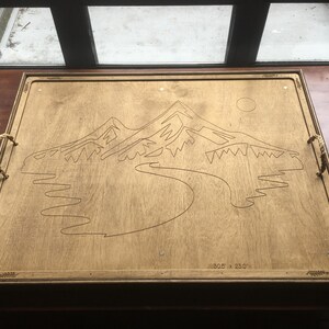 May include: Wooden serving tray with a mountain and river design. The tray has rope handles and measures 77.5 x 58.4 cm. The design is etched into the light-colored wood.