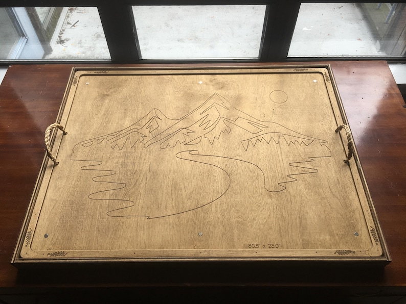 May include: Wooden serving tray with a mountain and river design. The tray has rope handles and measures 77.5 x 58.4 cm. The design is etched into the light-colored wood.