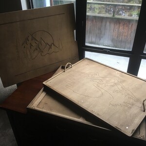 May include: Wooden serving trays with mountain and sun designs. The trays are light brown with etched artwork. One tray has rope handles. The trays are stacked on top of each other.