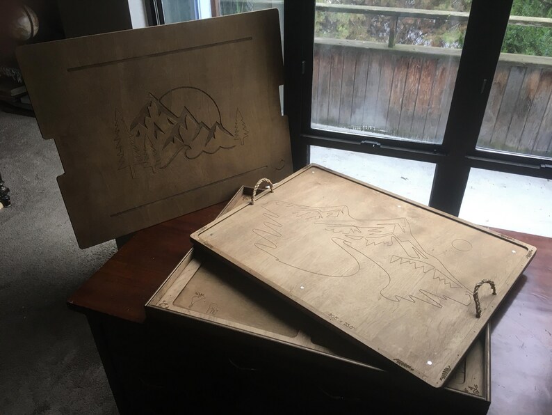 May include: Wooden serving trays with mountain and sun designs. The trays are light brown with etched artwork. One tray has rope handles. The trays are stacked on top of each other.