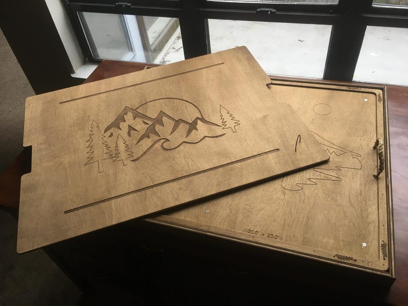 May include: Light brown wooden serving tray featuring a carved mountain and sun design. The tray has a landscape scene with mountains, trees, and a sun. Dimensions are 77.5 x 58.4 cm.