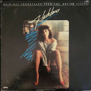 May include: A black vinyl record album cover with the title "Flashdance" in blue and white lettering. The cover features a woman wearing a gray sweater and red shoes, sitting on a wooden step with a brown carpet underneath.